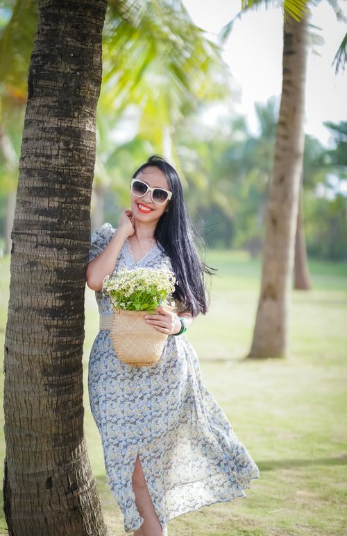 Free A stylish woman in sunglasses smiles holding flowers under palm trees in a sunny park. Stock Photo