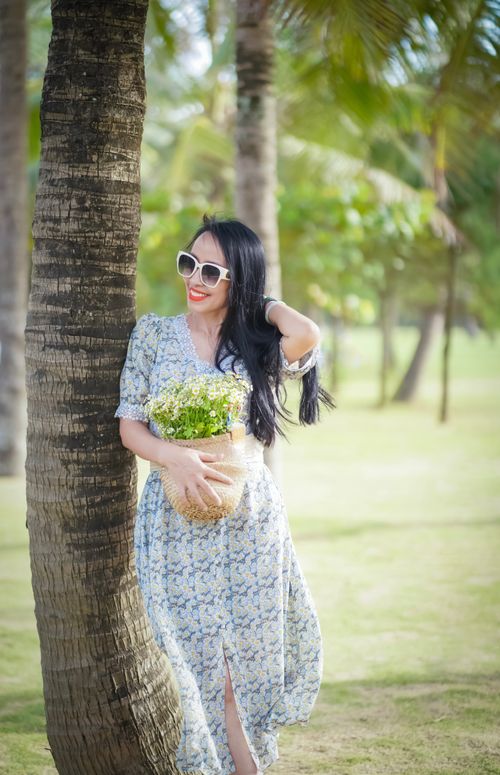 Free A woman elegantly posing in a summer dress, holding flowers and leaning against a palm tree outdoors. Stock Photo
