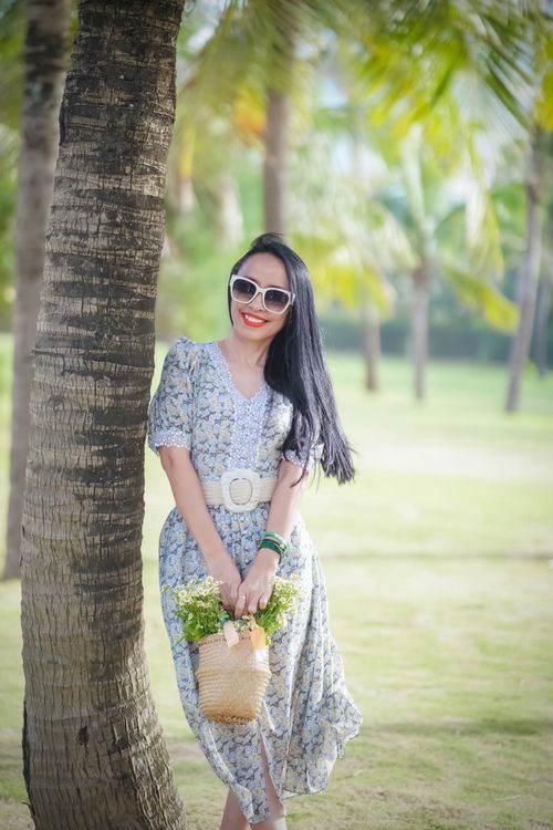 Free Smiling woman in floral dress leaning on a palm tree, enjoying a sunny day. Stock Photo
