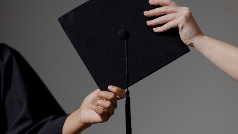 Two people exchanging a graduation cap on a grey background, symbolizing achievement.