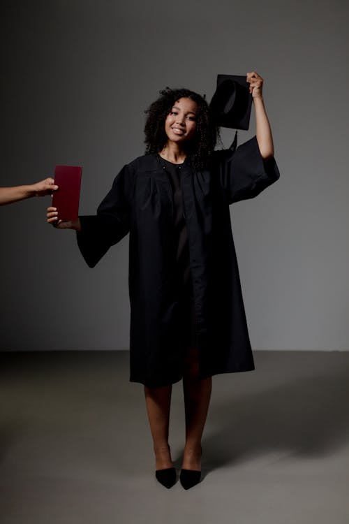 A Woman in Black Academic Regalia Smiling while Holding Her Mortarboard