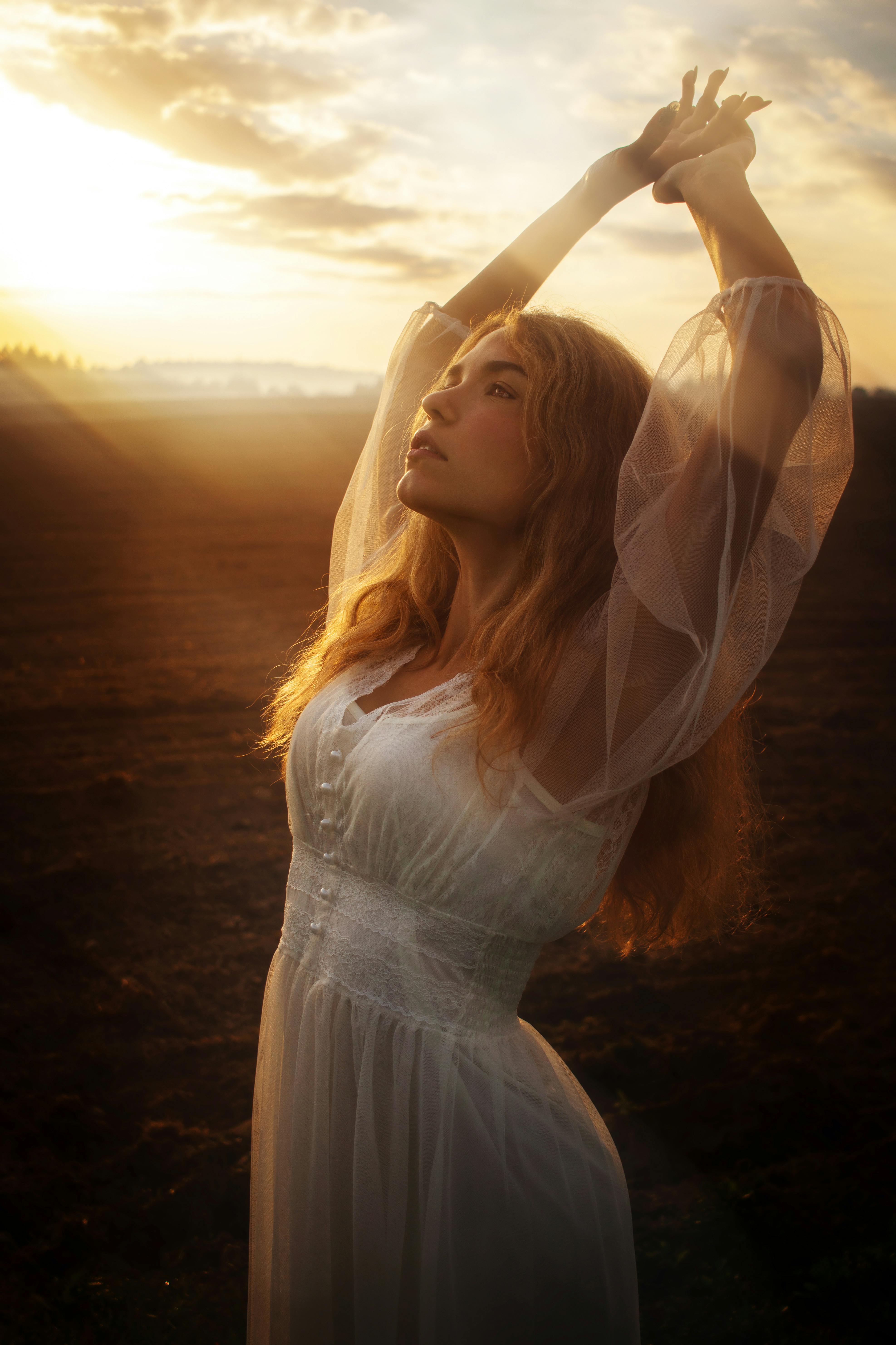 A woman in a flowing dress stands elegantly in a rural field during sunset.