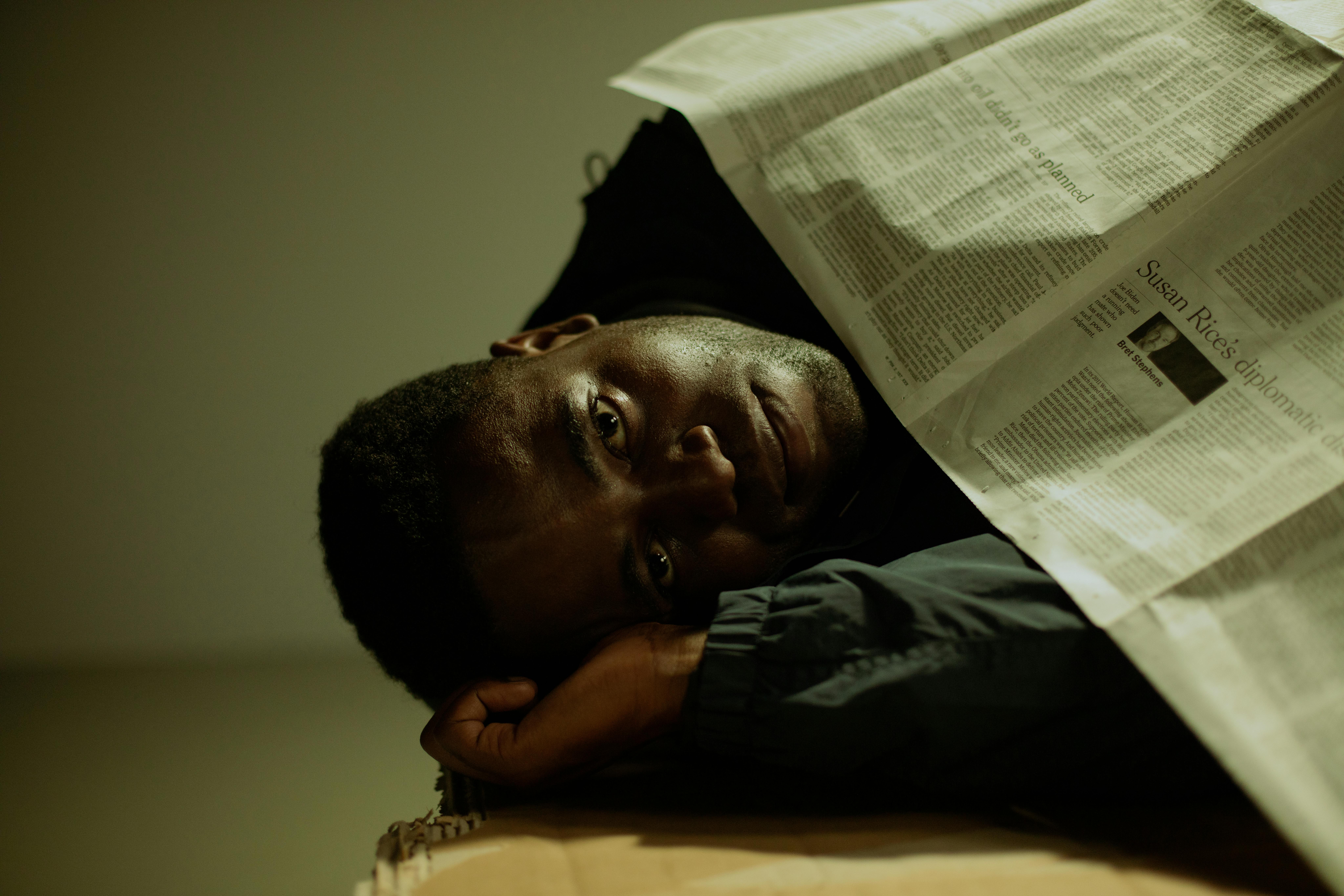 A man rests indoors, covered with a newspaper, symbolizing themes of homelessness and poverty.