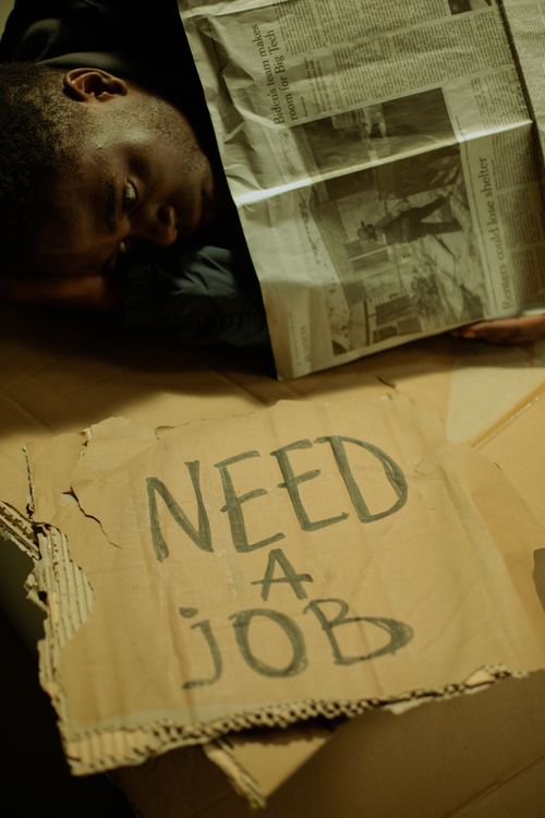 Free A man lies on cardboard indoors with a 'Need a Job' sign, highlighting social issues. Stock Photo