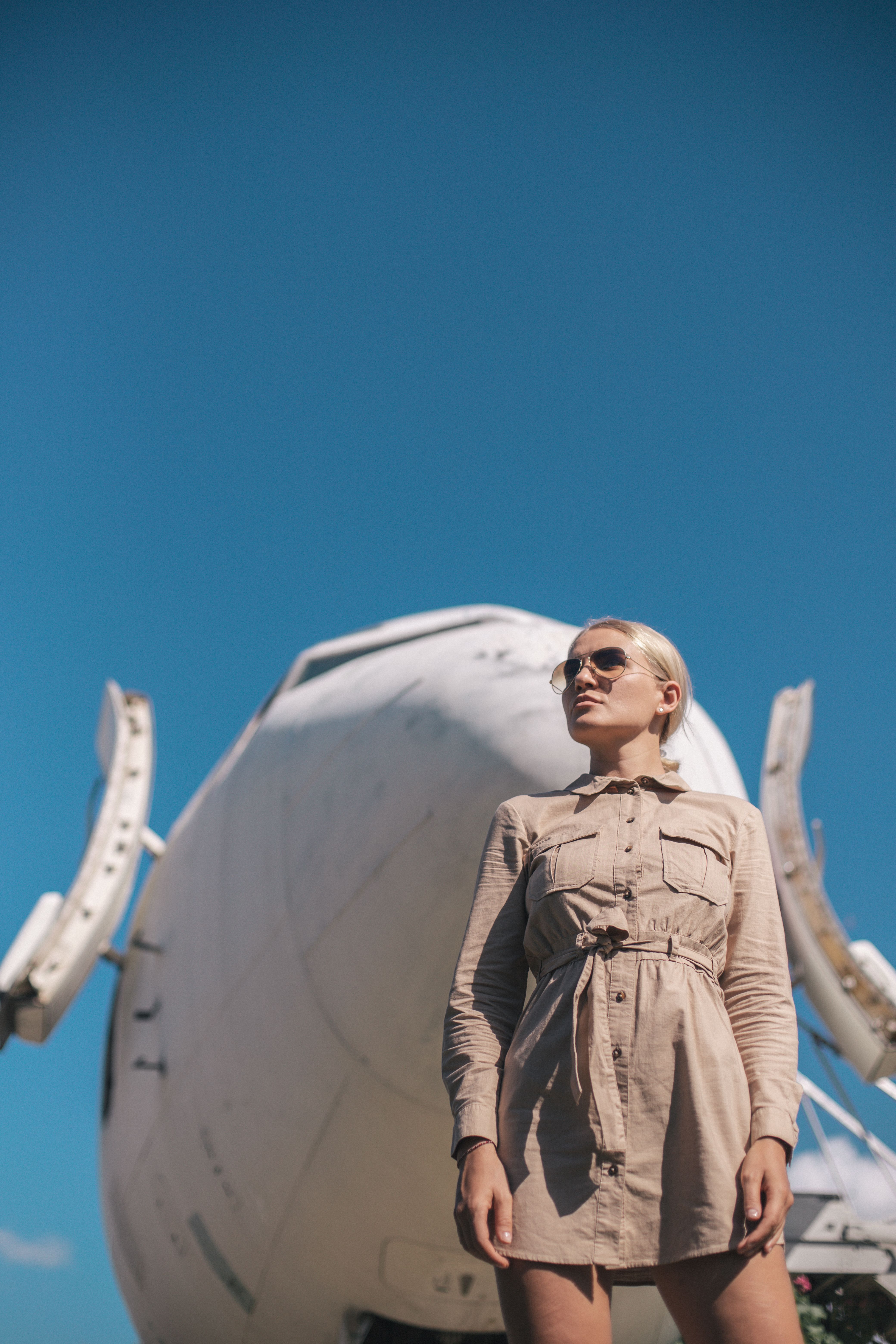 Confident woman poses in front of airplane on a sunny day with clear blue sky.
