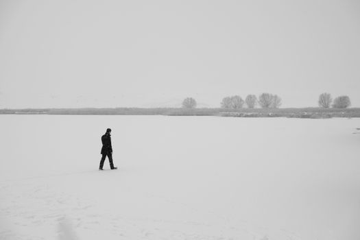 A lone figure in black walks across a vast, snowy landscape on a winter day.
