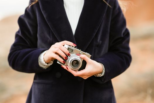 A stylish woman with manicured nails holds a vintage camera while wearing a black jacket outdoors.