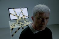 An elderly man looking at camera and computer tied with a black and yellow tape behind him