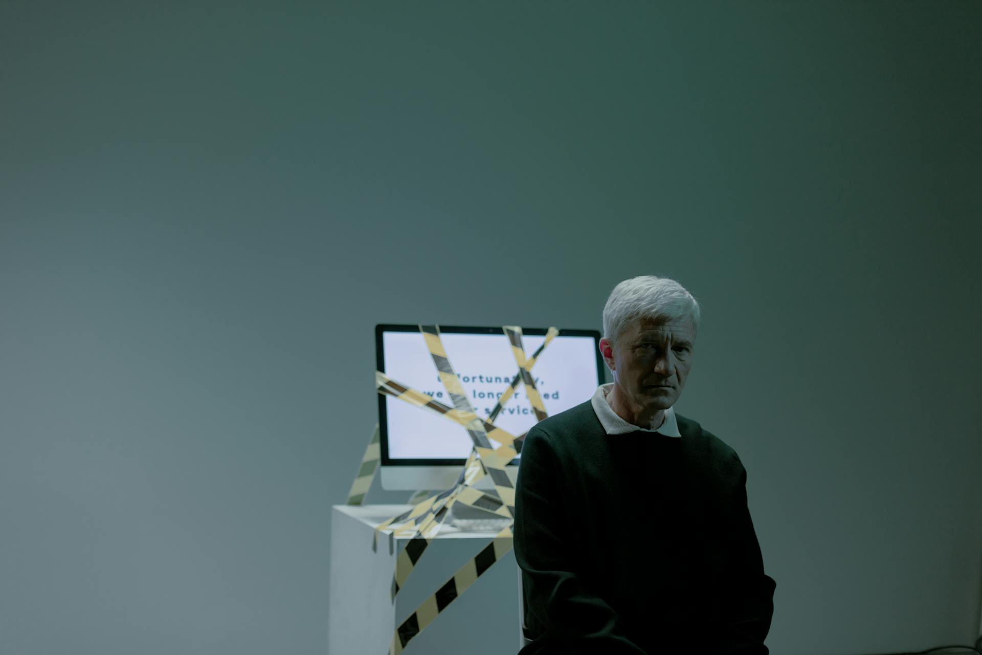 A senior adult sits indoors next to a computer wrapped in caution tape, symbolizing job loss and unemployment.