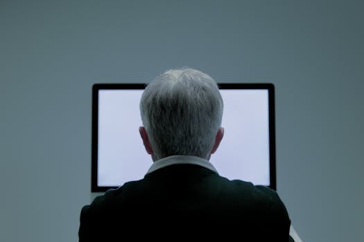 Rear view of a senior man with grey hair using a computer screen indoors.