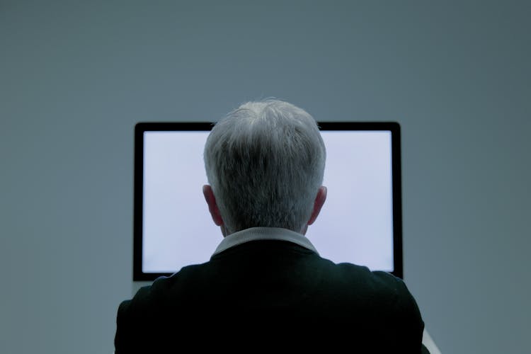 Elderly Man Sitting In Front Of A Computer Seen From Behind