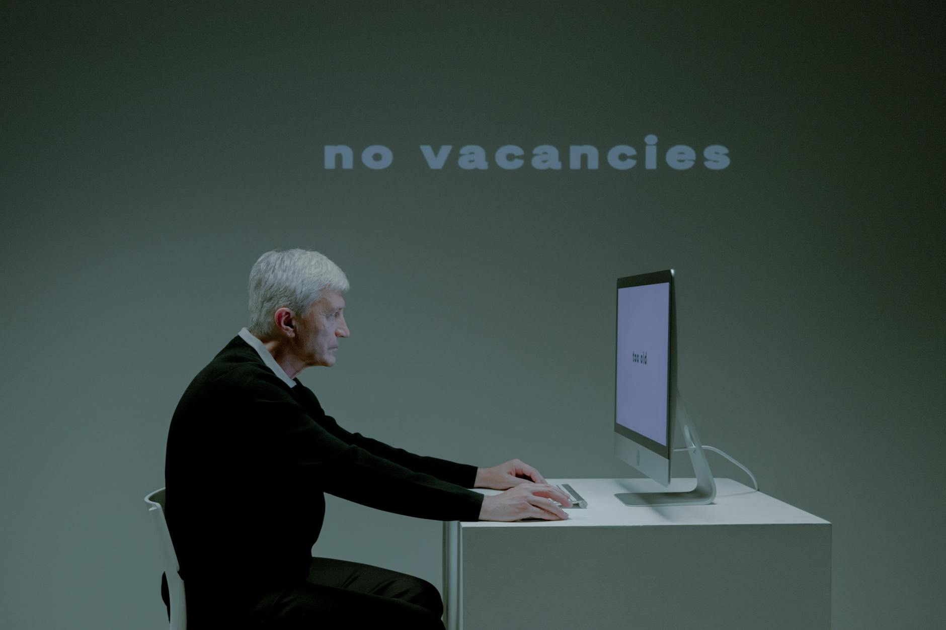 Senior man sitting at a computer with 'no vacancies' displayed, highlighting unemployment issues.