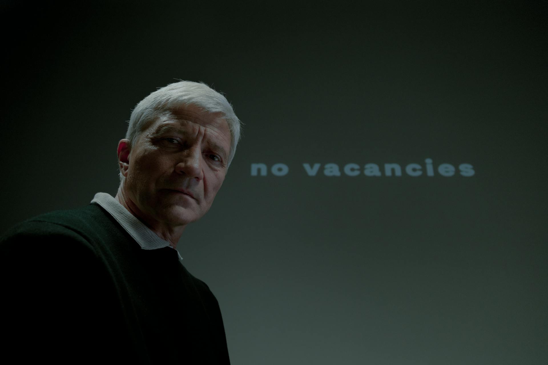 Senior man looks serious in front of a no vacancies message highlighting unemployment issues.