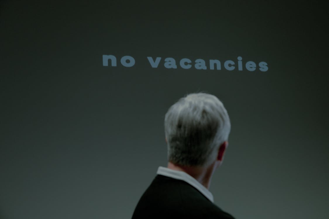 Man with gray hair looking at a projected “no vacancies” sign on a dark background.