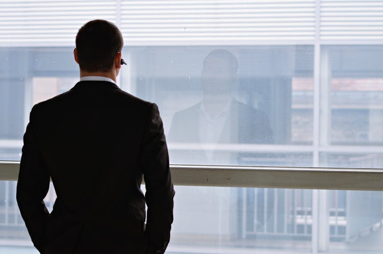 A Back View Of A Man In Black Suit Facing The Glass Window