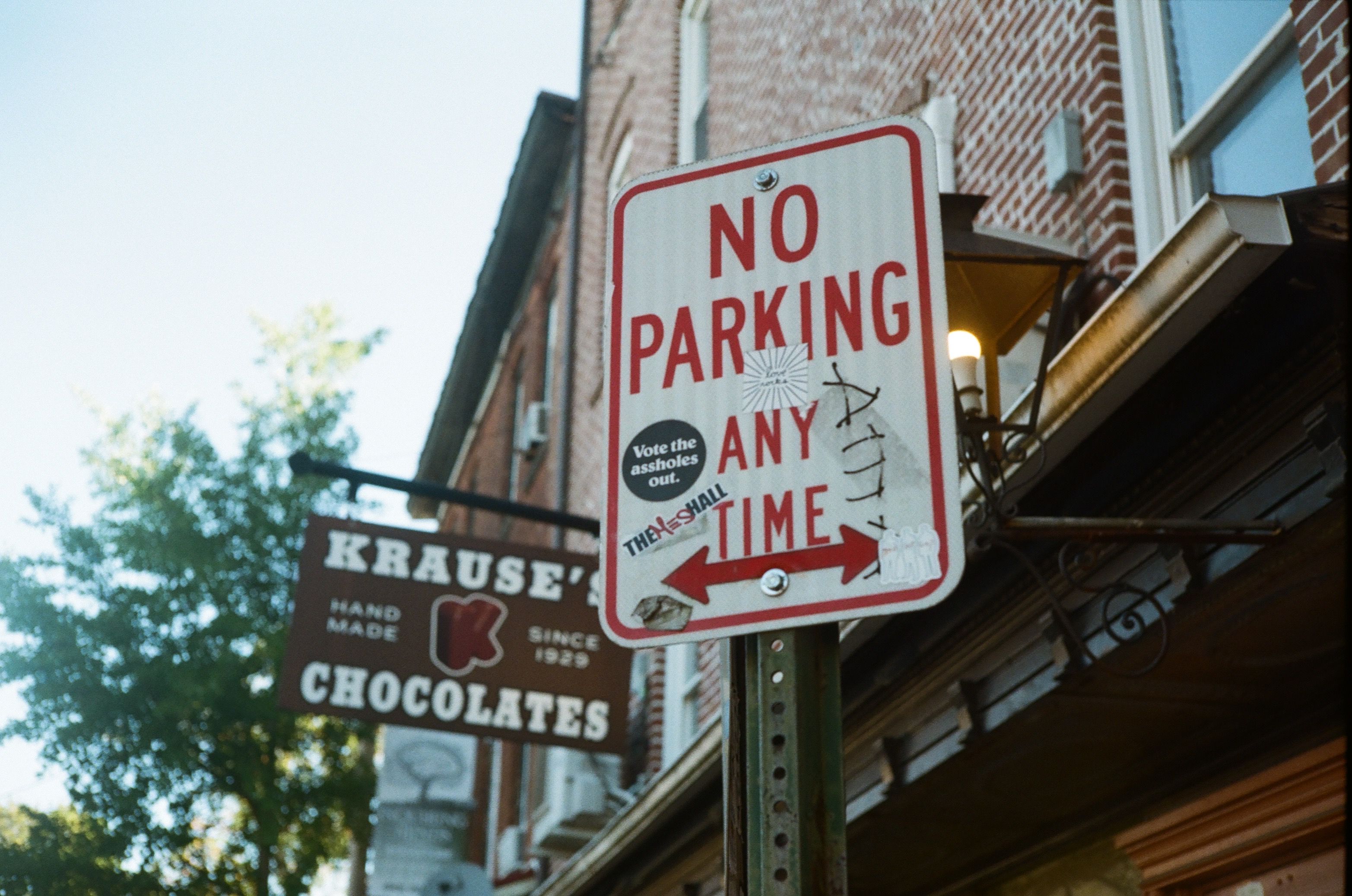 Close-up of a 'No Parking' sign on a city street with a chocolate shop sign in the background.
