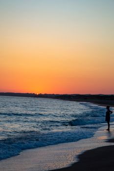 A lone figure stands at the water's edge, silhouetted against a vibrant sunset over the ocean.