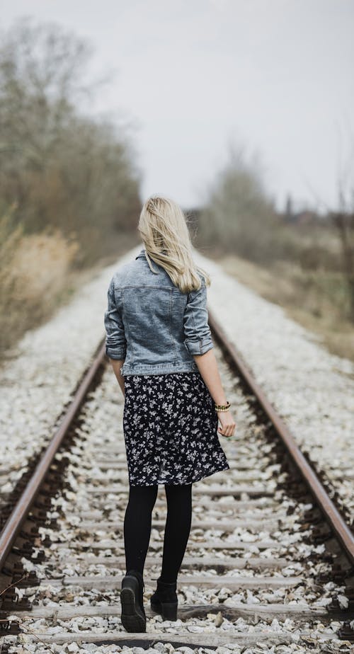 Mujer En Chaqueta De Mezclilla Azul, Polainas Negras Y Vestido Blanco Y Negro Caminando Sobre Riel De Tren