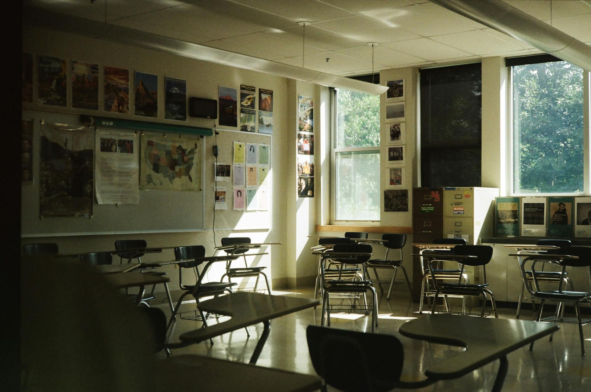 A sunlit classroom with empty desks, chairs, and educational posters on the walls.