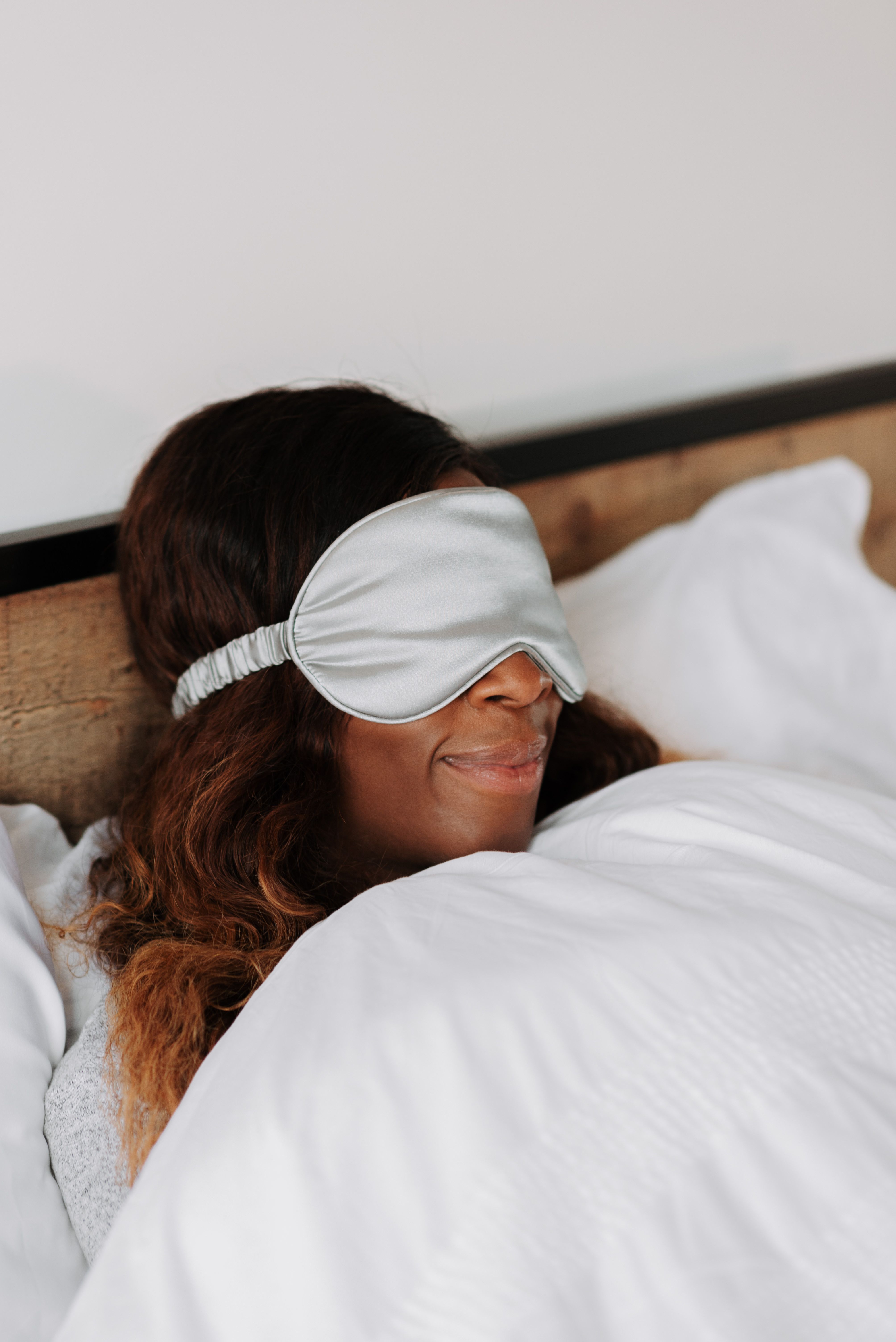 Woman resting in bed under a white duvet wearing a silver satin sleep mask