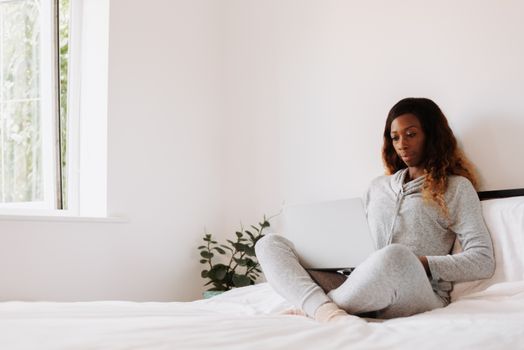 Woman sitting cross-legged on a bed using a laptop. Bright, cozy bedroom atmosphere.
