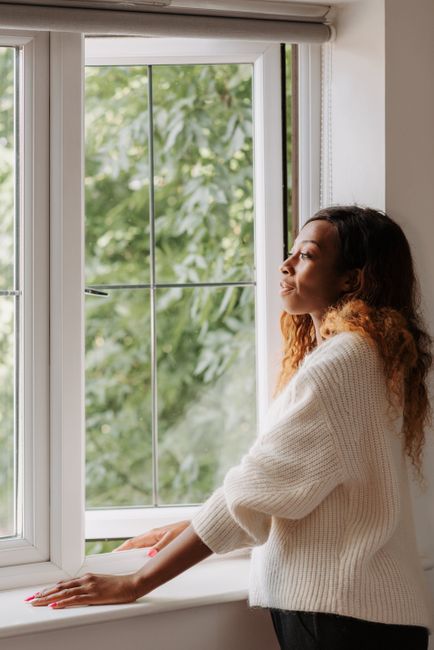 A woman in a white sweater gazes out of a window, enjoying a peaceful moment indoors.