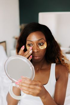 Woman with gold eye patches looking in mirror, focused on skincare.