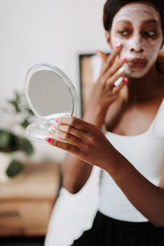 A woman applies a white face mask while holding a handheld mirror in a cozy bedroom setting.