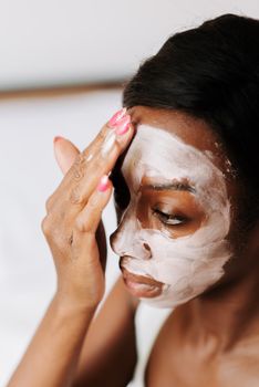 A woman with curly hair applies a face mask for skincare in a close-up indoor shot.