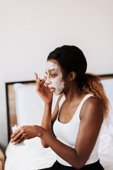 A young woman with a skincare face mask sits in her bedroom, focusing on her beauty routine.