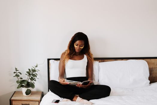 Woman with under-eye patches reading a magazine in a cozy bedroom setting.
