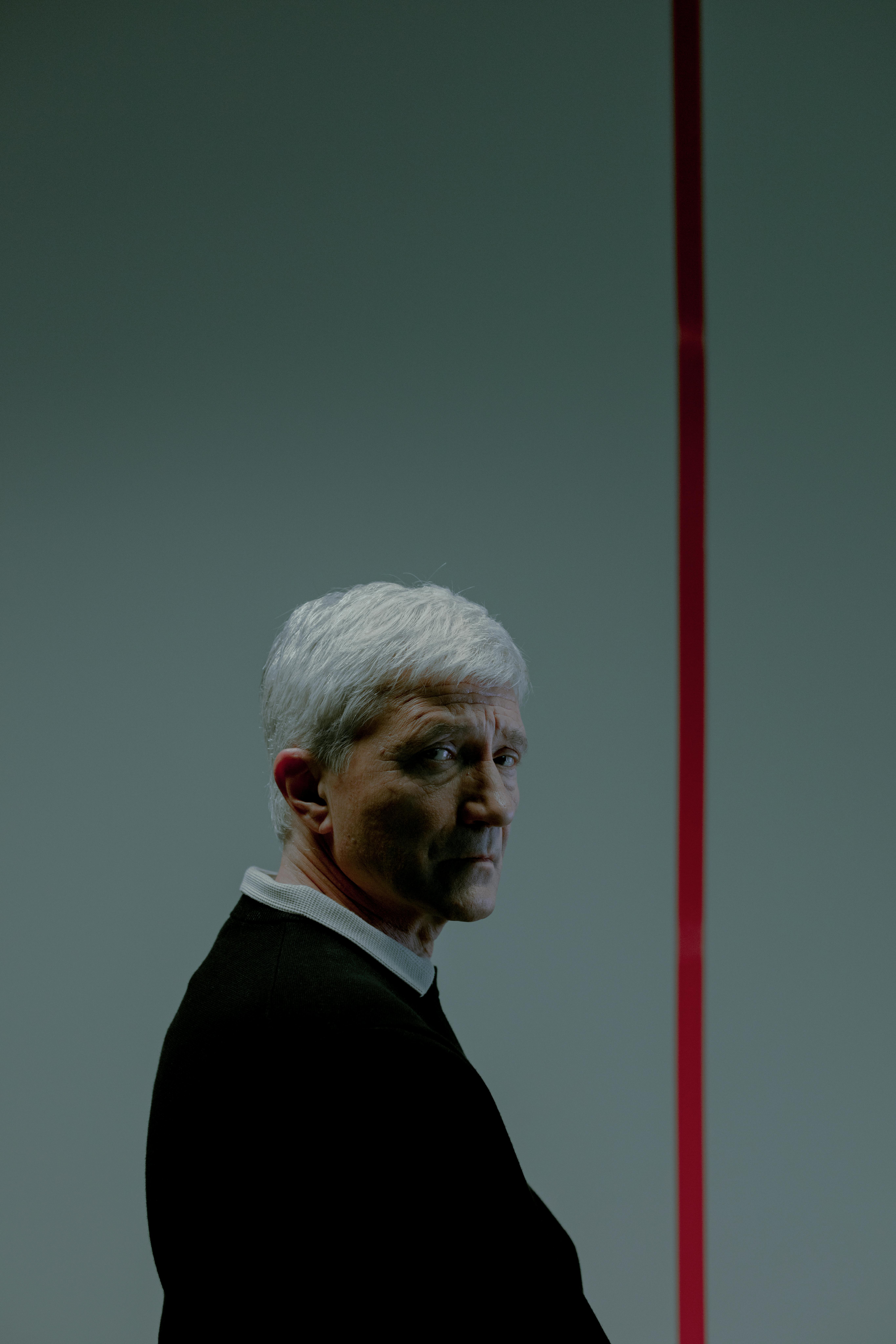 A thoughtful senior man in a dark sweater with white hair stands indoors, facing sideways.