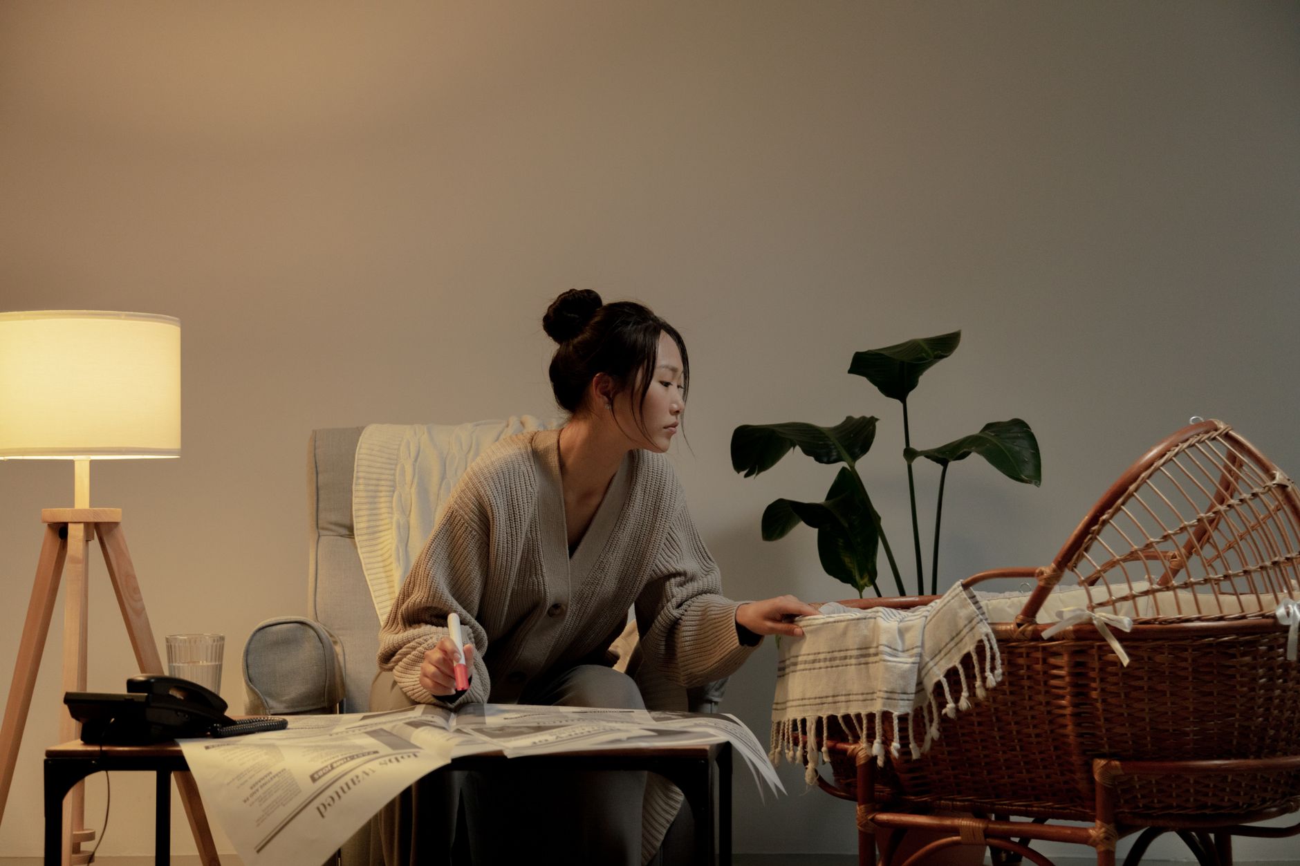 A woman multitasking in a living room, reading a newspaper and tending to a baby in a stroller.