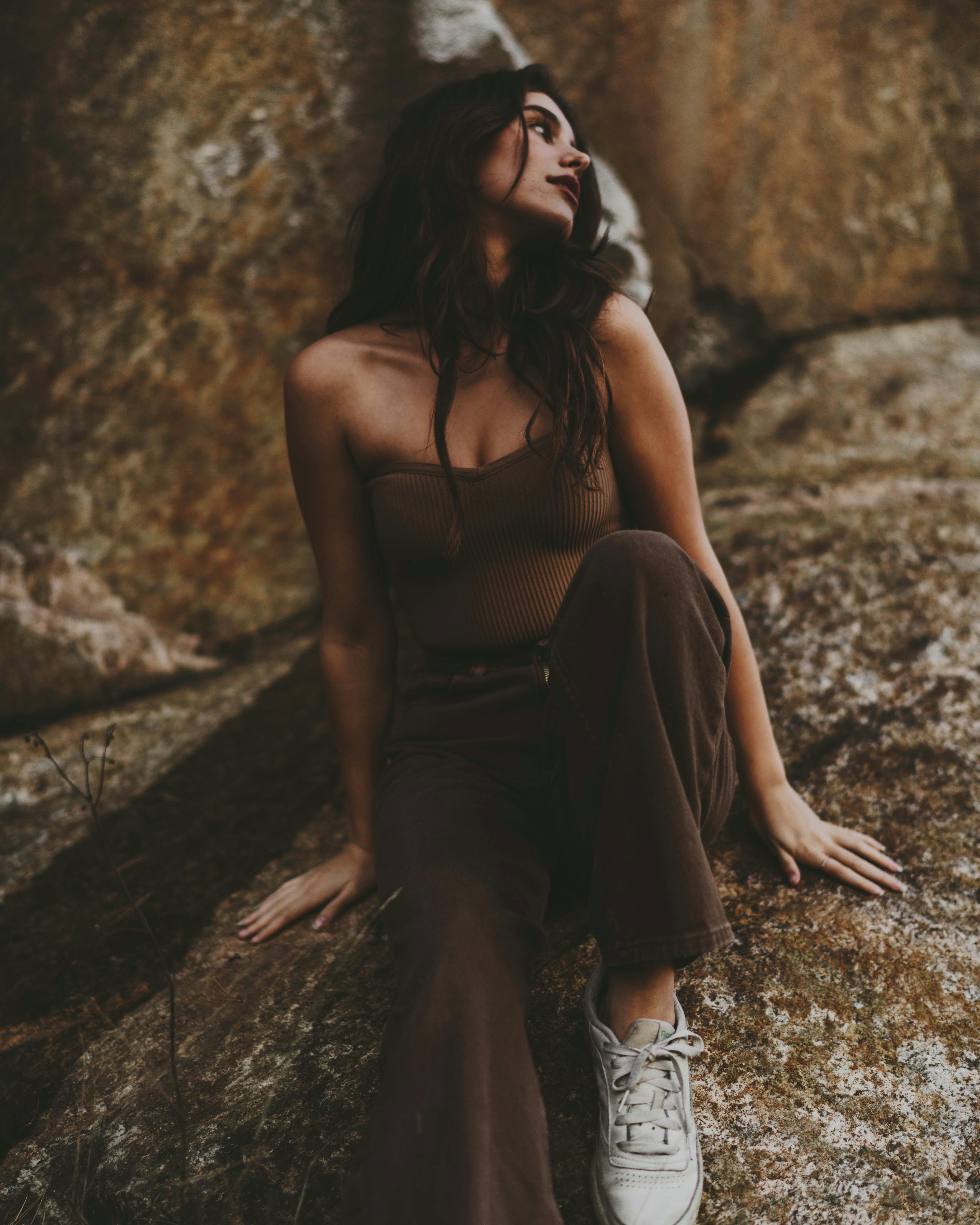 Woman sitting on a rock, wearing brown pants and tube top, in natural setting.