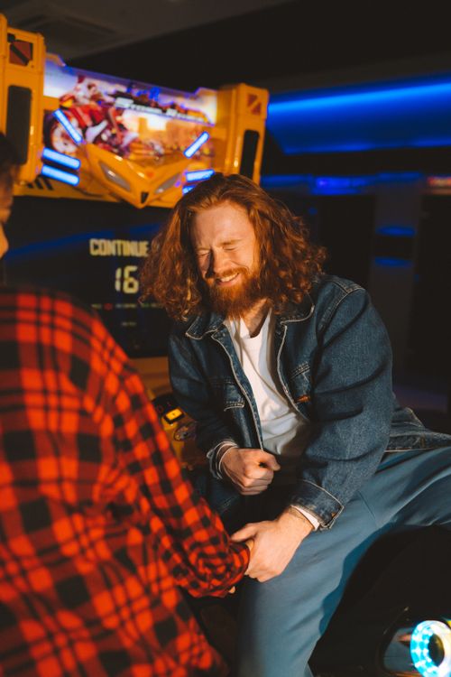 Free Man with long hair and beard laughing in an arcade, wearing a denim jacket. Fun moment captured. Stock Photo