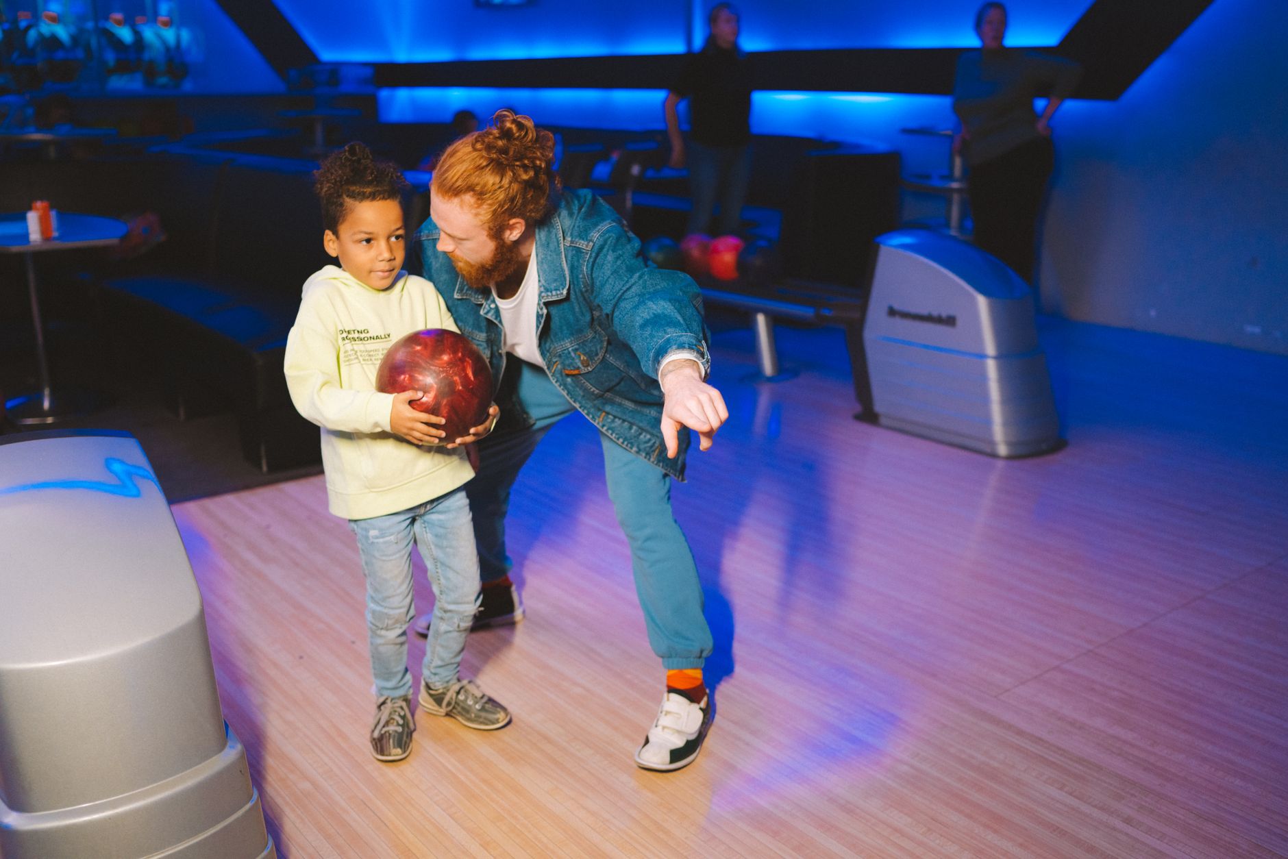 A bonding moment as a father teaches his young son to bowl at an indoor alley under blue lights.