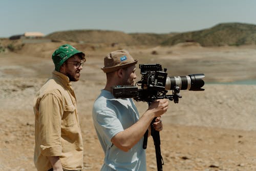 A Man in Brown Bucket Hat Holding a Video Camera while Standing Beside the Man in Green Bucket Hat