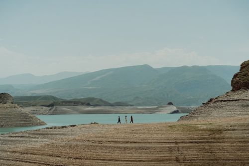 Free Three people walk along a tranquil lake surrounded by mountains. Stock Photo