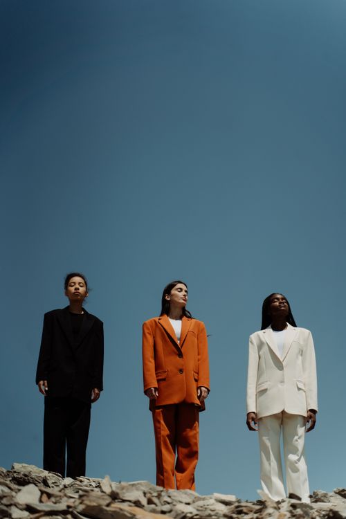 Free Three women wearing fashionable blazers stand confidently outdoors against a clear blue sky. Stock Photo