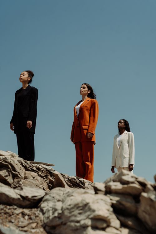 Free Three women in diverse suits pose confidently on rocky terrain against a clear blue sky. Stock Photo