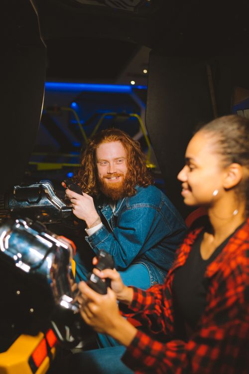 Free Diverse couple playing a racing game at an arcade, sharing smiles and competitive spirit. Stock Photo