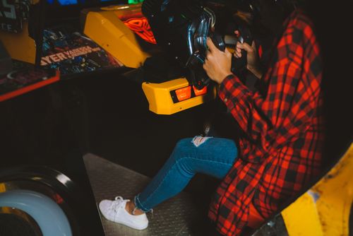 Free Young woman enjoying an arcade racing game wearing a checkered shirt. Fun and leisure activity captured indoors. Stock Photo