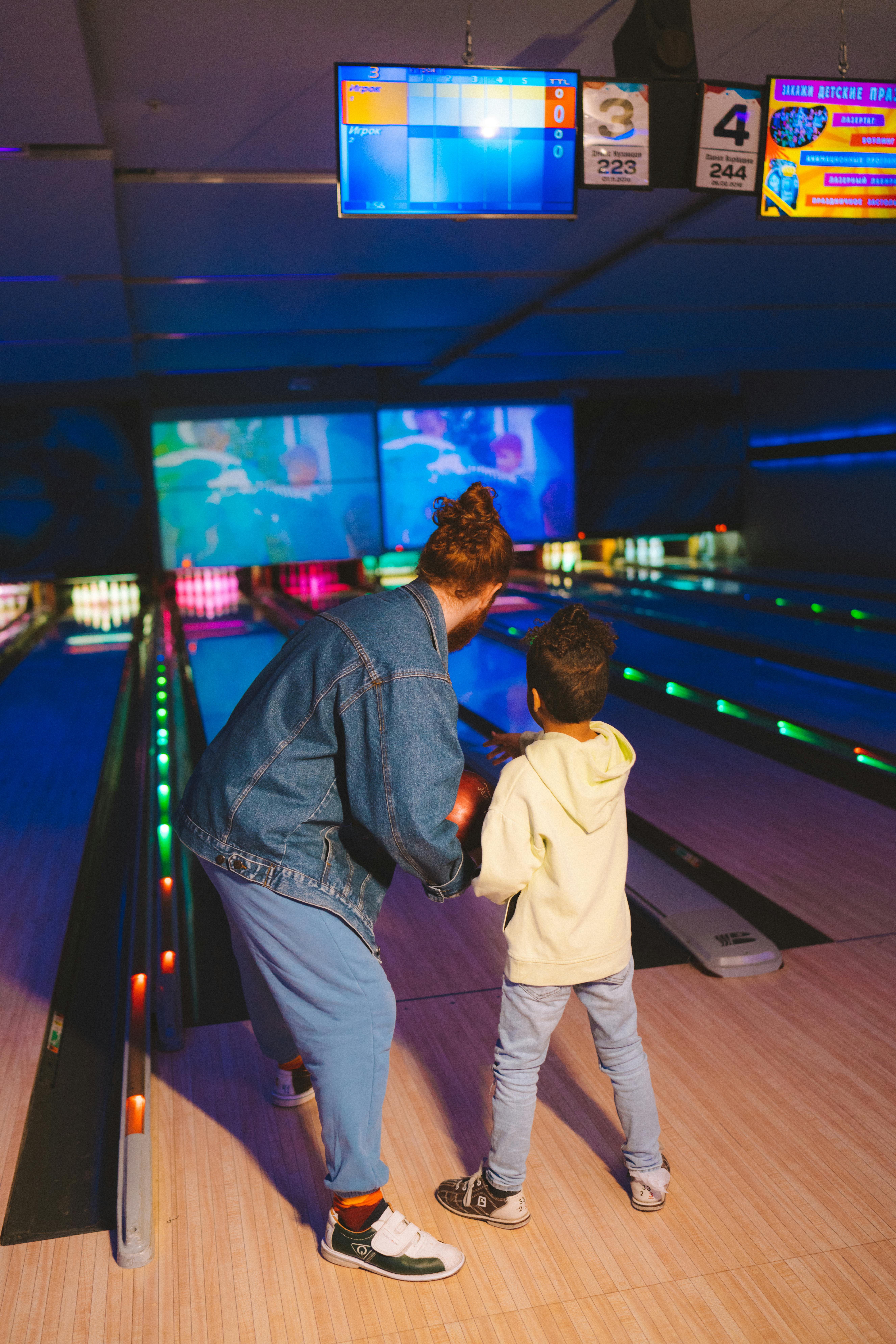 Woman and child bowling, preparing to bowl. They stand at the lane; screens above display scores.