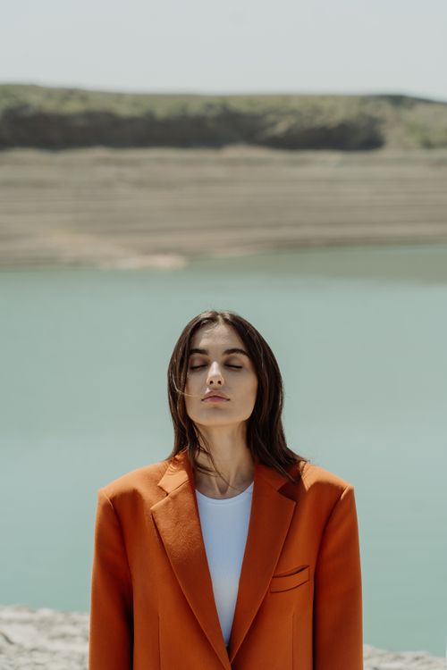 Free Portrait of a woman in an orange coat with closed eyes, standing by a lake. Stock Photo