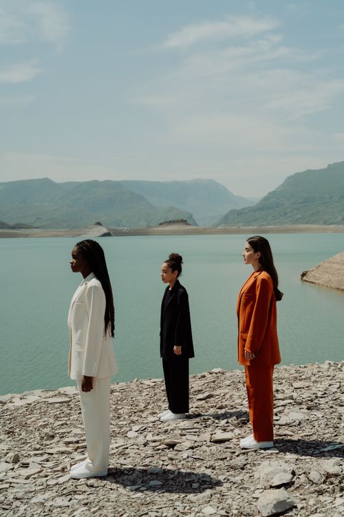 Free Three diverse women in business attire stand on a cliffside by a serene lake, symbolizing unity and empowerment. Stock Photo