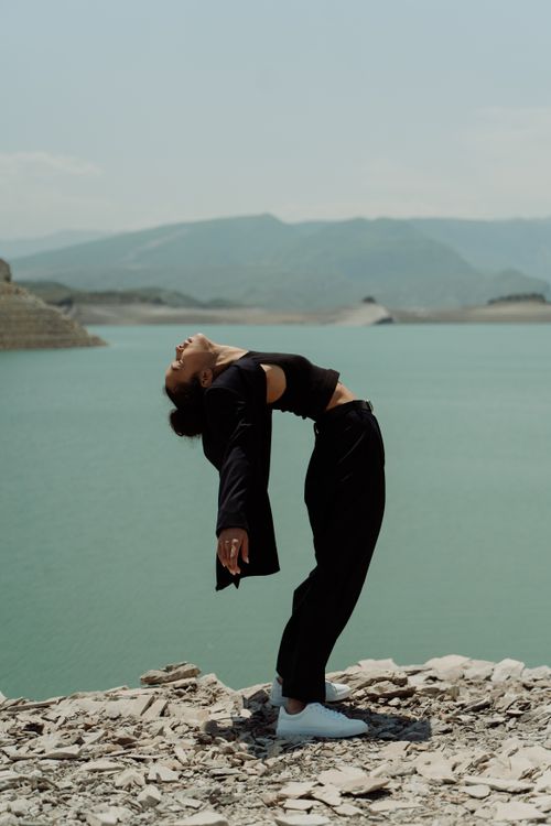 Free An Asian woman in black clothing artfully balances on a rocky lakeside with mountains in the background. Stock Photo