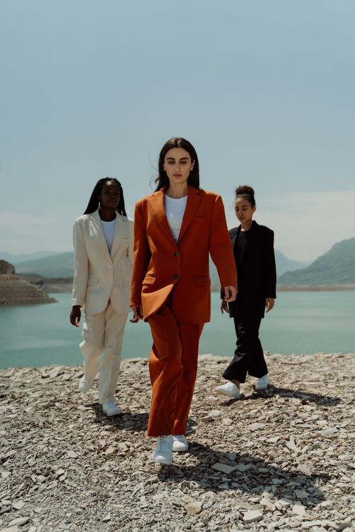 Free Three diverse women in suits walk confidently on a rocky landscape by a lake. Stock Photo