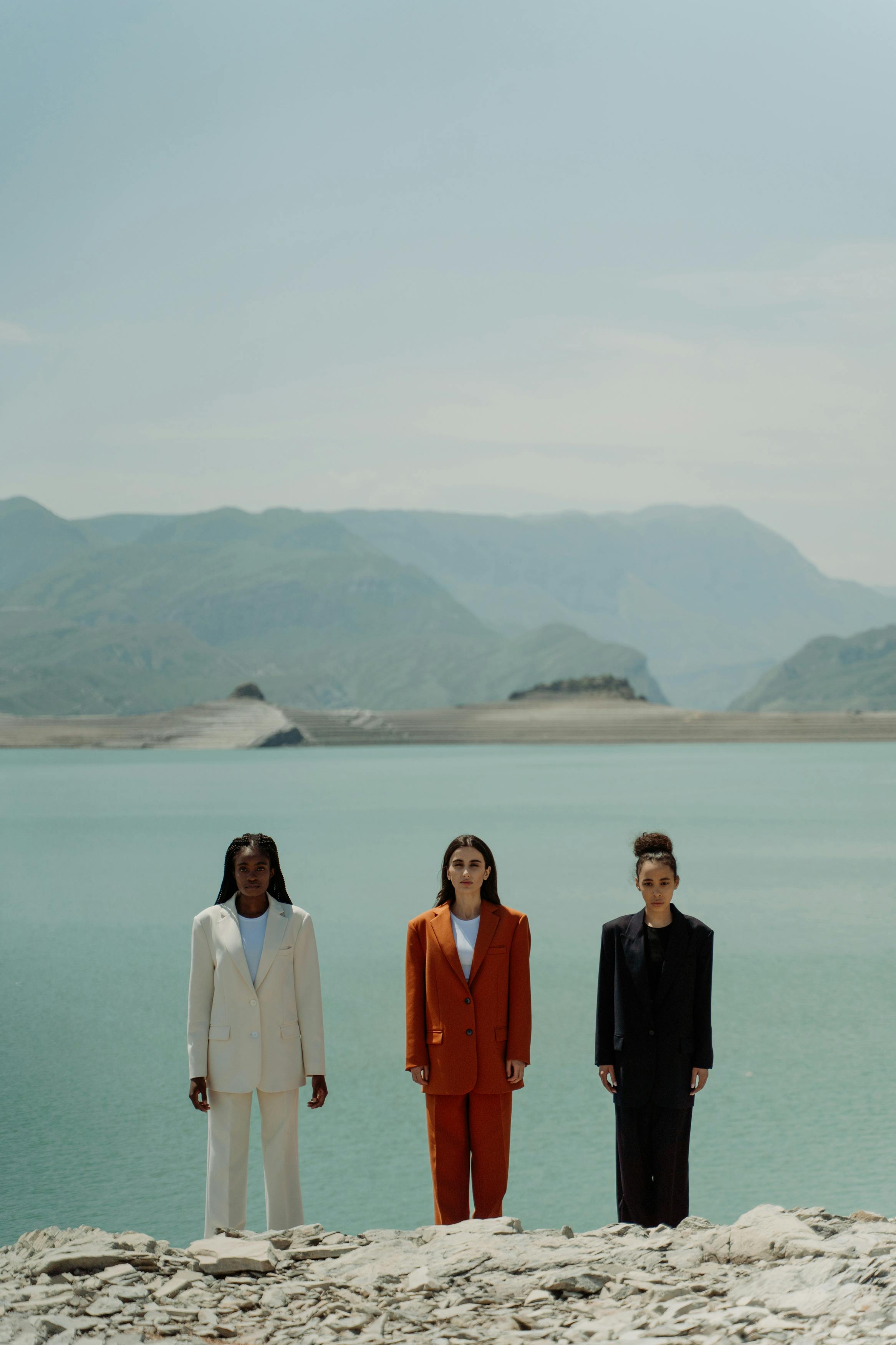 Three diverse women in stylish suits posing by a serene mountain lake under a clear sky.