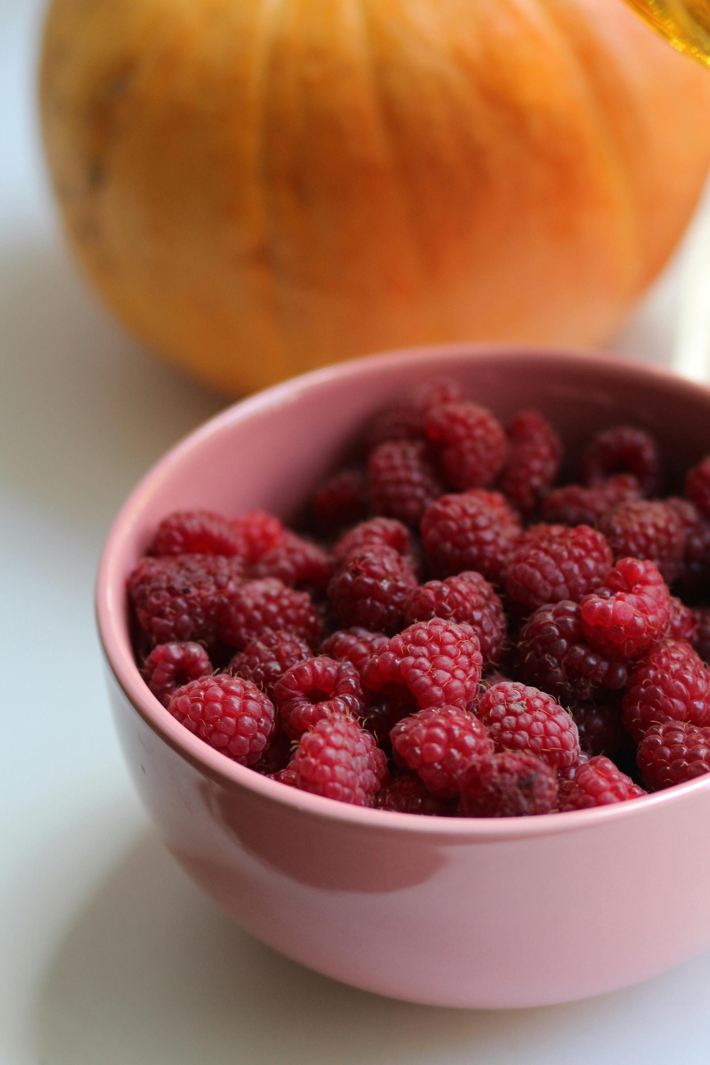 A pink bowl filled with fresh raspberries next to a melon on a light background.