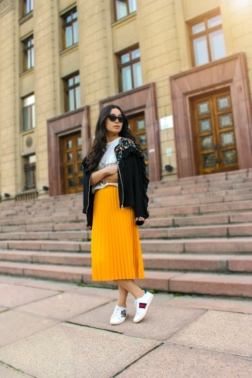 Free Women Wearing Black Jacket and Pleated Yellow Skirt Standing on Brown Floor Stock Photo